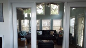 A living room with a black leather sofa, a coffee table, and a dining table. There are two windows with blinds, a ceiling fan, and a plant in the corner.