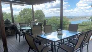 A covered outdoor dining area with a glass table and chairs, overlooking a lake and trees.