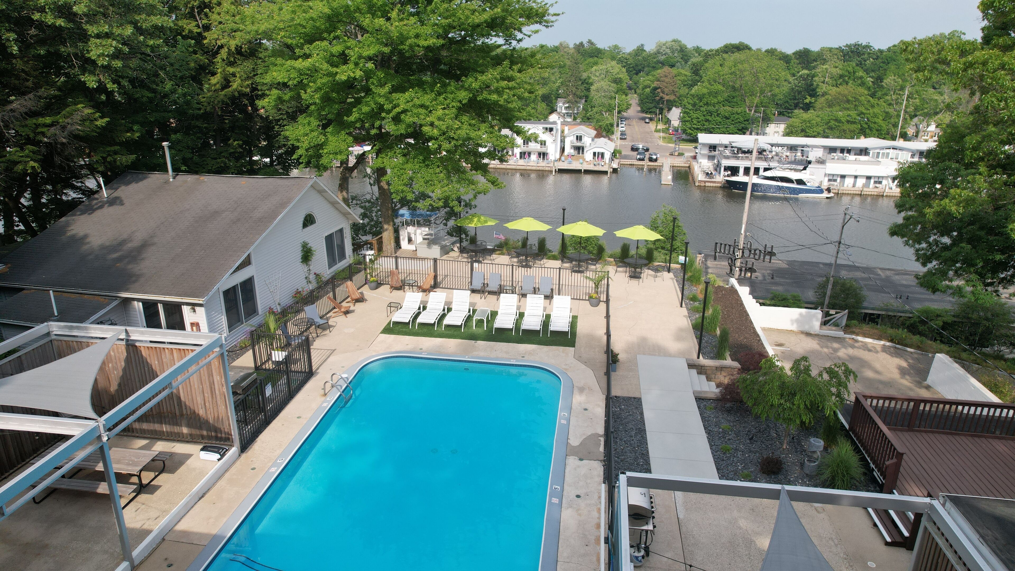 A swimming pool with lounge chairs and umbrellas, a dock with boats, and a view of a lake and buildings in the background.
