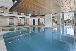 Una piscina interior moderna con agua cristalina, techo de madera y paredes de vidrio que ofrecen vistas de la ciudad.