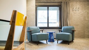 Ein Zimmer mit zwei Sesseln, einem kleinen blauen Tisch, einem großen Fenster mit Vorhängen und einer Ziegelwand.