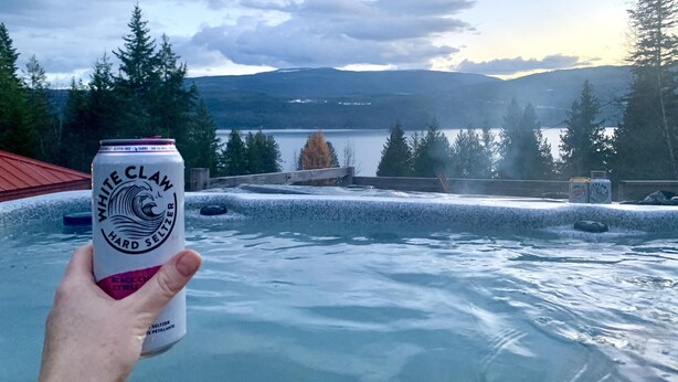Una mano sosteniendo una lata de White Claw Hard Seltzer frente a un jacuzzi con una vista panorámica de montañas y árboles.