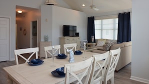 A dining area with a table set for four, a television in the background, and a living room with a sofa and curtains. - Rehoboth Beach Condo Elevator Pool Parking Clean (Rehoboth Beach)