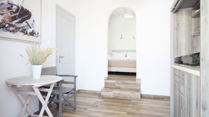 A modern kitchen with wooden flooring, a dining area with a table and chairs, and a bedroom with a bed and an arched doorway. - Crystal View Mykonos (Mykonos)