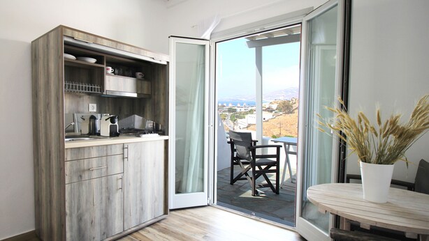 A modern kitchen with a wooden cabinet, a dining area with a table and chairs, and a sliding glass door leading to a balcony with a view. - Crystal View Mykonos (Mykonos)