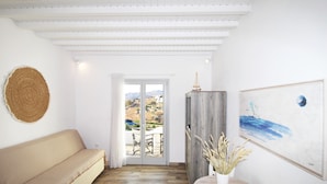 A room with a beige sofa, a wooden floor, a large painting, and a view through a glass door. - Crystal View Mykonos (Mykonos)