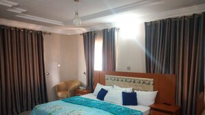 A hotel room with a bed, a chair, a nightstand, and curtains. - Bayse One Place Jericho (Ibadan)