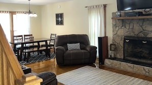 A spacious living room with a high ceiling, a stone fireplace, a large flat-screen TV, and a dining area with a wooden table and chairs.
