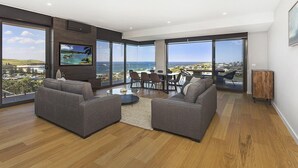 A spacious living room with a large TV, grey sofas, a coffee table, and a dining area with chairs. - Cosmopolitan@SoHo -  amazing views (Gerringong)