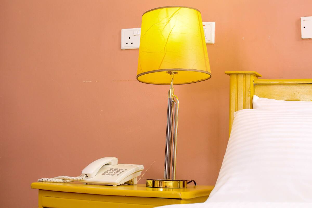 A bed with white linens, a yellow bedside table with a white telephone, and a yellow lamp with a yellow shade.