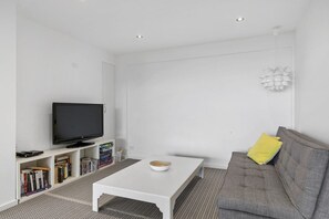 A modern living room with a flat-screen TV, a white coffee table, a grey sofa, and a bookshelf with books.