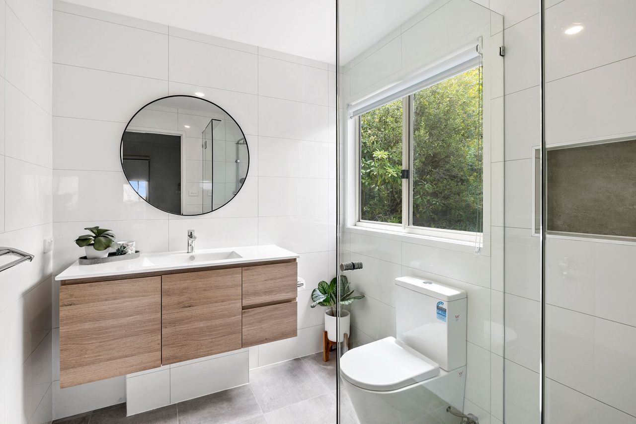 A modern bathroom with a white vanity, a round mirror, a toilet, and a window with a view of greenery.