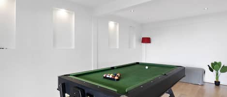 A pool table with balls in the center of a room with white walls and wooden flooring.