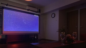 Une salle de cinéma privée avec un grand écran diffusant une image de ciel étoilé, une horloge au mur et une table ornée de verres à vin et de fleurs.