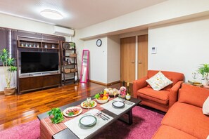 A living room with a flat-screen TV, wooden entertainment unit, orange sofa, dining table set with plates, glasses, and a bottle of wine. - Mizu House (Naha)