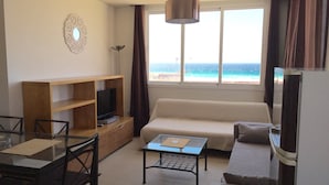 A living room with a sofa, a coffee table, a TV on a wooden stand, a dining table with chairs, and a view of the ocean through large windows. - SENSES BUILDING HAPPINESS APARTMENT (Tarifa)