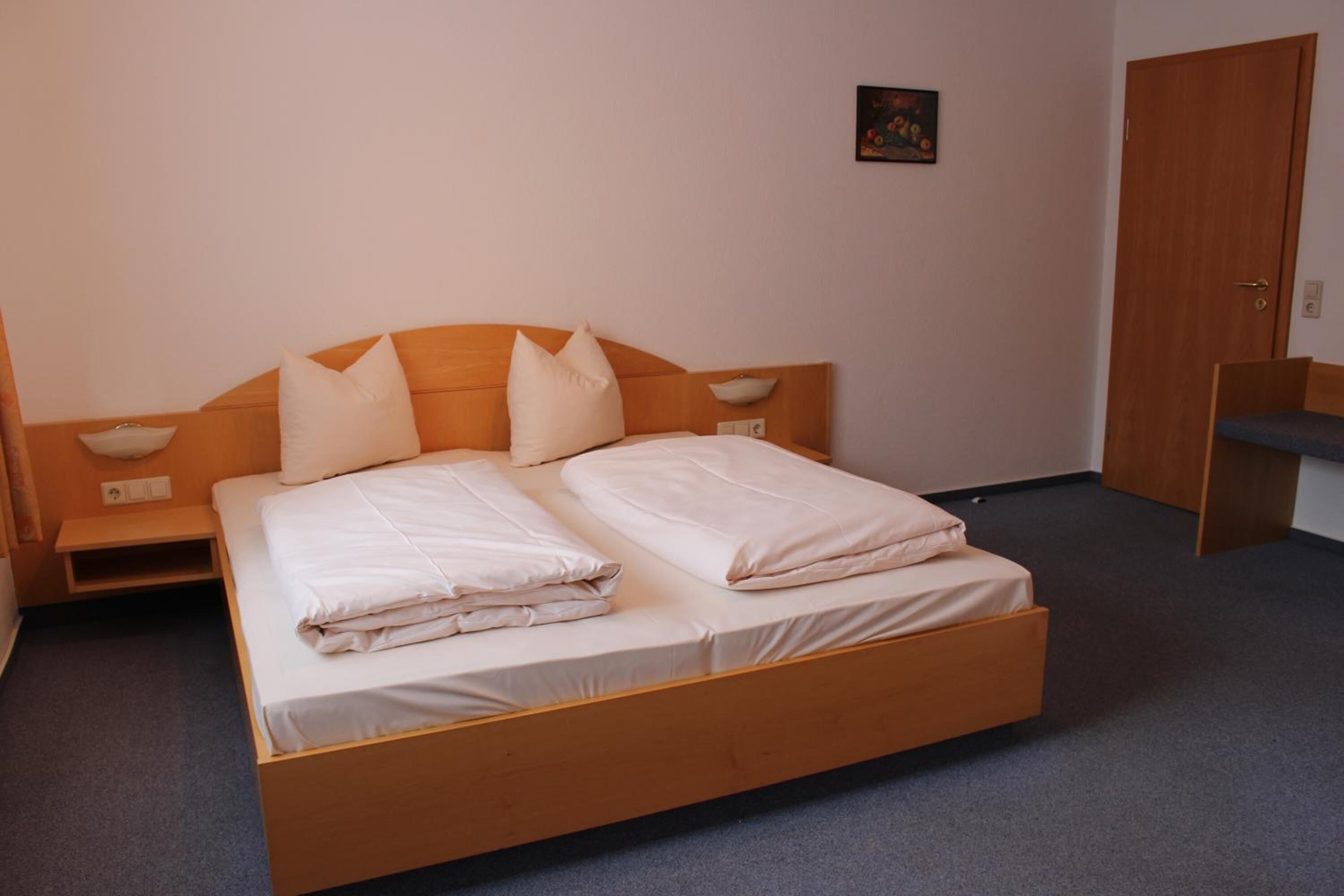A single bed with a wooden headboard, two pillows, and a folded blanket. There is a bedside table with a lamp, a small shelf, and a wall-mounted light. A door is visible to the right.