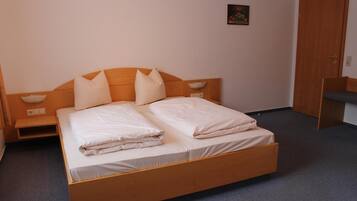 A single bed with a wooden headboard, two pillows, and a folded blanket. There is a bedside table with a lamp, a small shelf, and a wall-mounted light. A door is visible to the right.