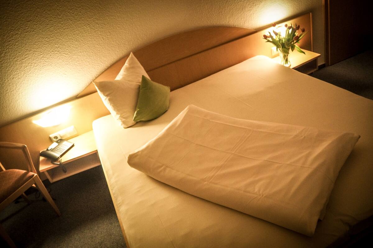 Ein ordentlich bezogenes Bett mit Kissen, ein Nachttisch mit Lampe und ein Sessel.