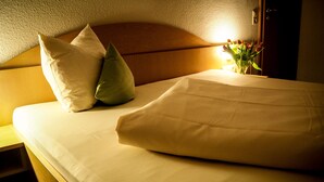 Una cama bien tendida con sábanas blancas y dos almohadas, una mesita de noche y un jarrón con flores.