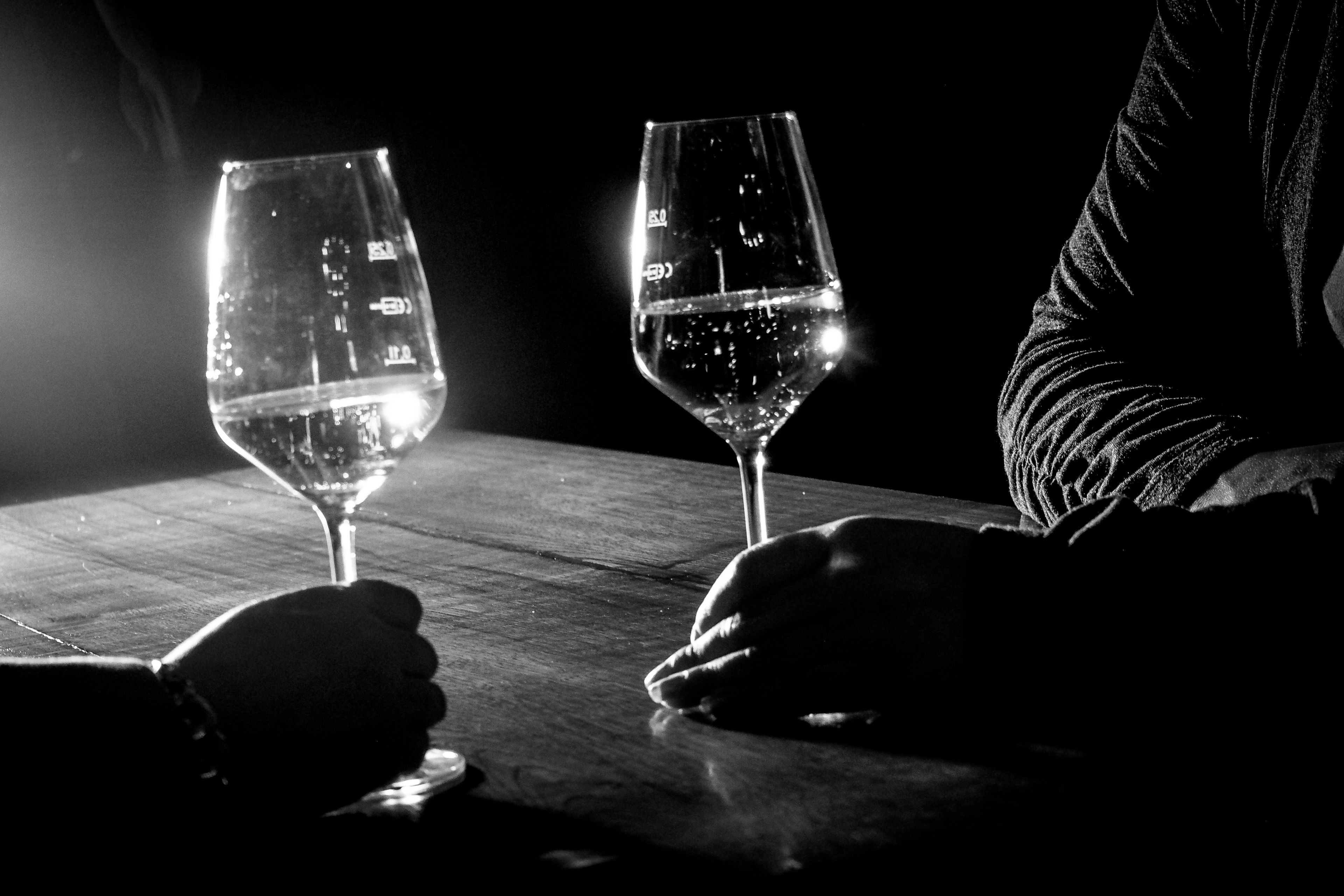 Two wine glasses on a wooden table with a person's hands resting on them.