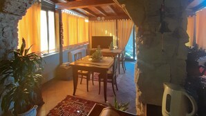 Un comedor acogedor con un arco de piedra, mesas y sillas de madera. A la izquierda hay una planta grande en maceta y a la derecha, un hervidor.