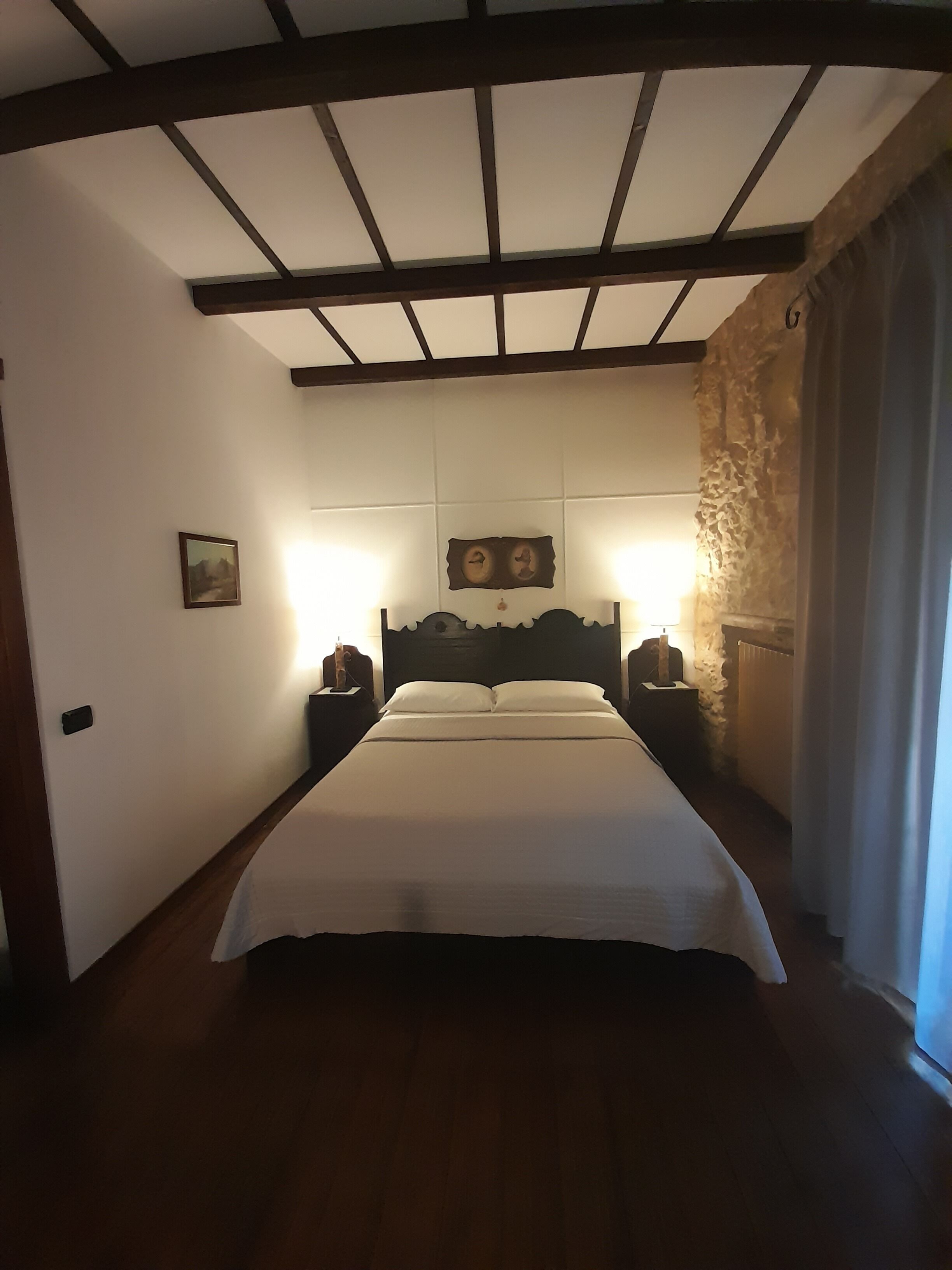A bedroom with a large bed, wooden beams on the ceiling, a stone wall, and a nightstand with a lamp.