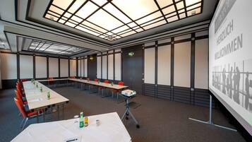 Un salón de conferencias con una larga mesa, sillas, un proyector y una pantalla mostrando 'HERZLICH WELCOME'.