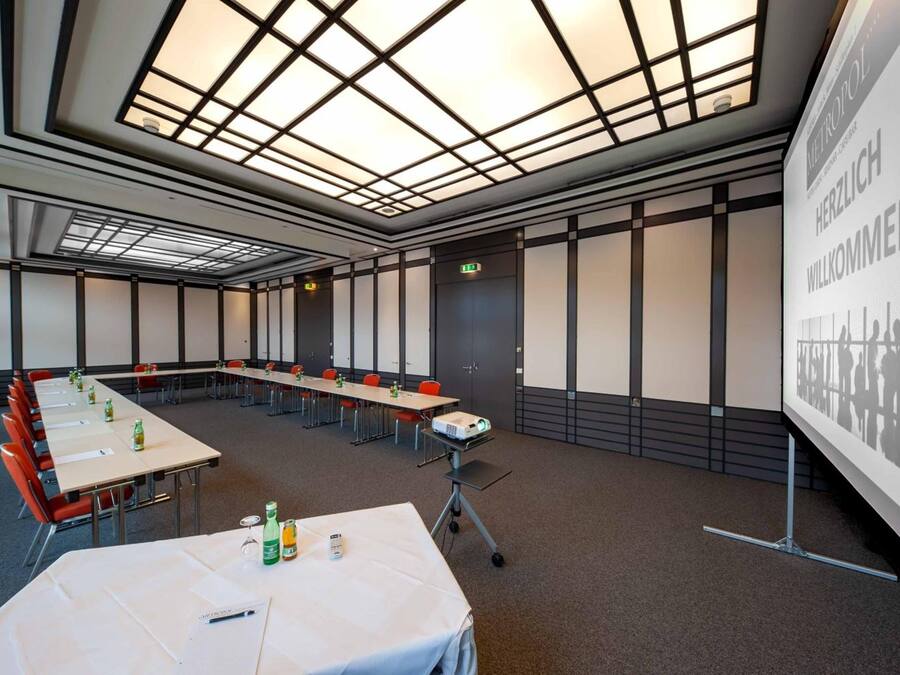 A conference room with a long table, chairs, a projector, and a screen displaying 'HERZLICH WELCOME'.