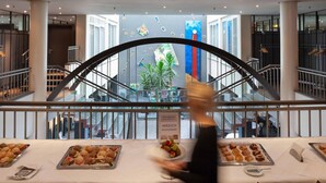 Zona de bufé con diversas opciones de comida, incluyendo sándwiches y pasteles, en un moderno entorno hotelero con vistas al vestíbulo y la escalera.