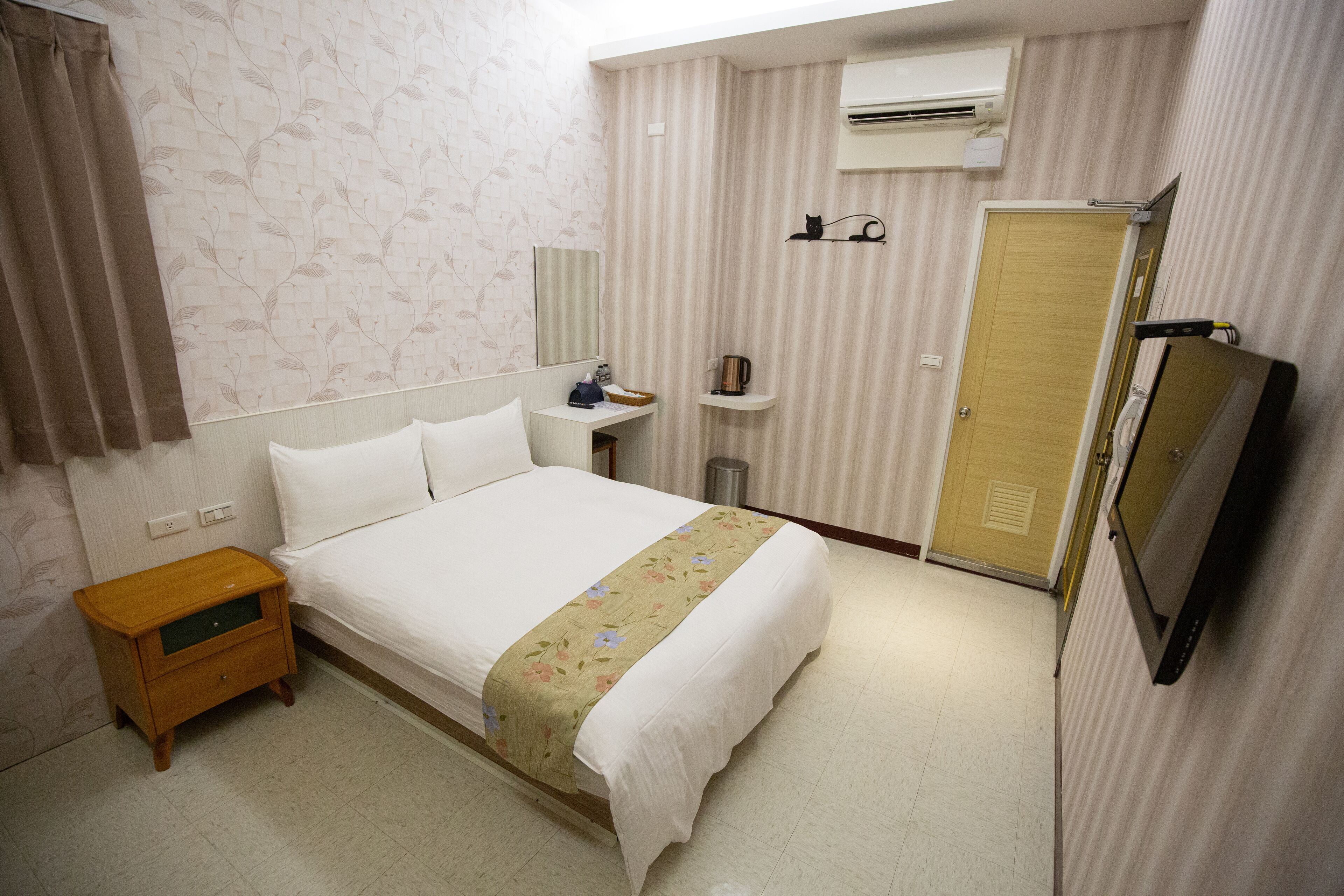 A hotel room with a bed, a nightstand, a TV, a wardrobe, and an air conditioning unit.