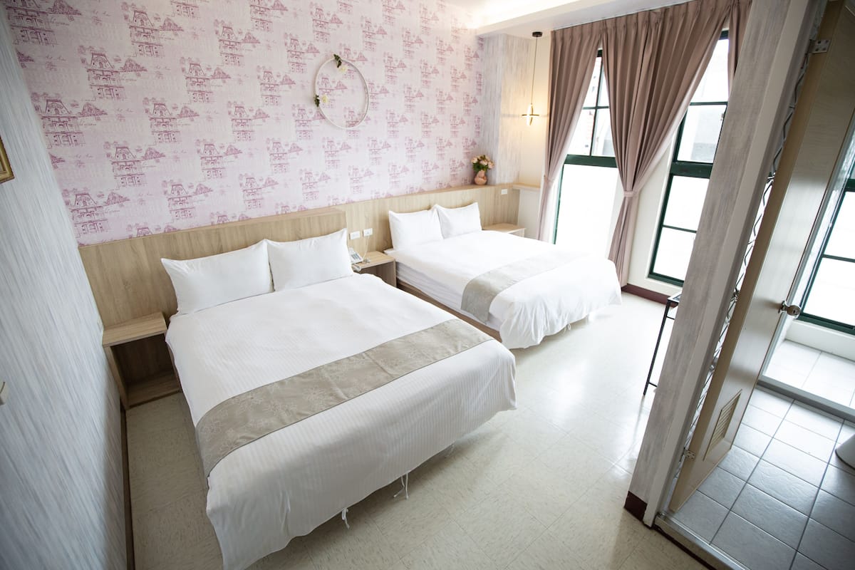 A hotel room with two beds, a wooden headboard, a wall with a pink pattern, and a window with curtains.