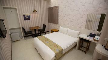 A hotel room with a bed, bedside table, chair, desk, and a small stool.