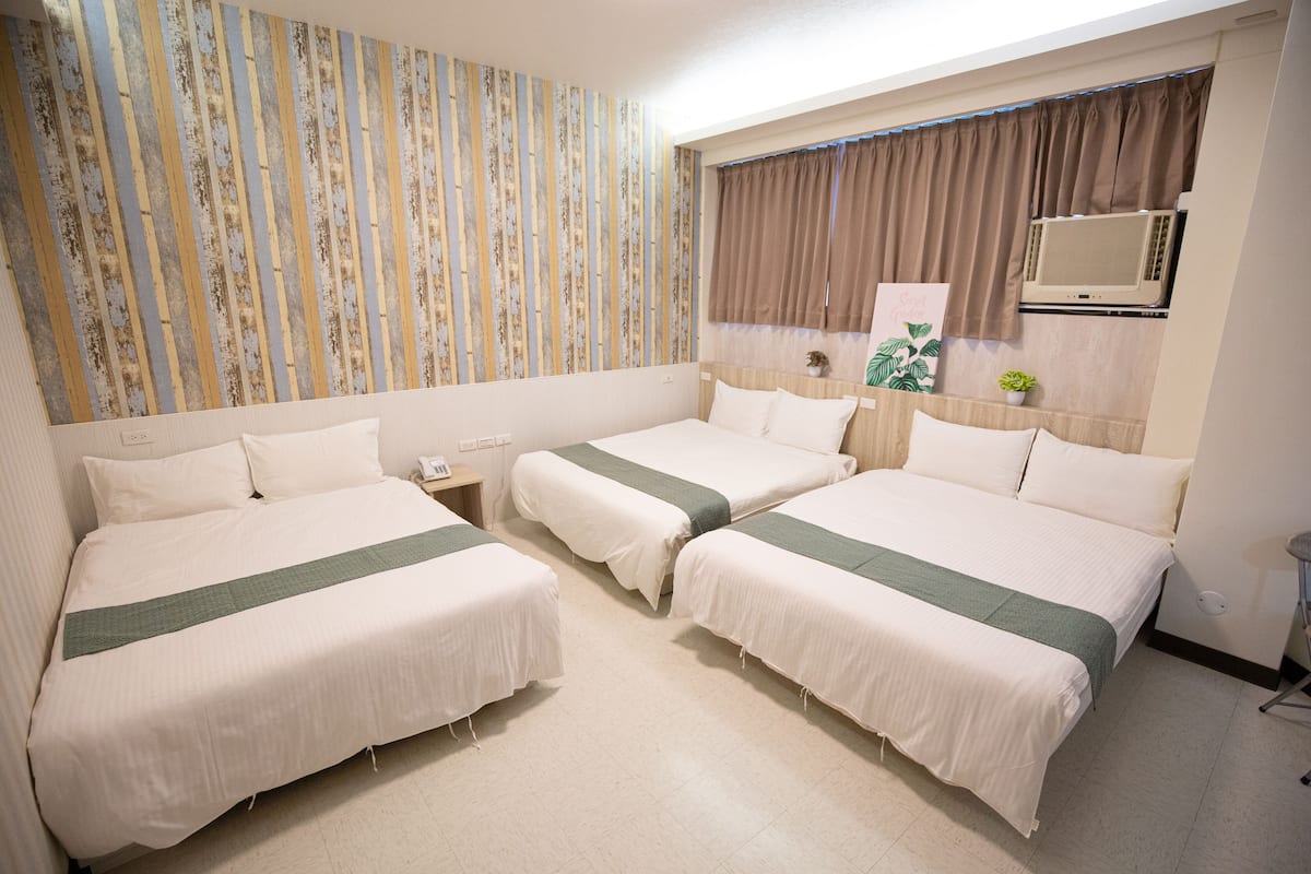 A hotel room with three single beds, a large window with curtains, an air conditioning unit, and a wall with a striped pattern.