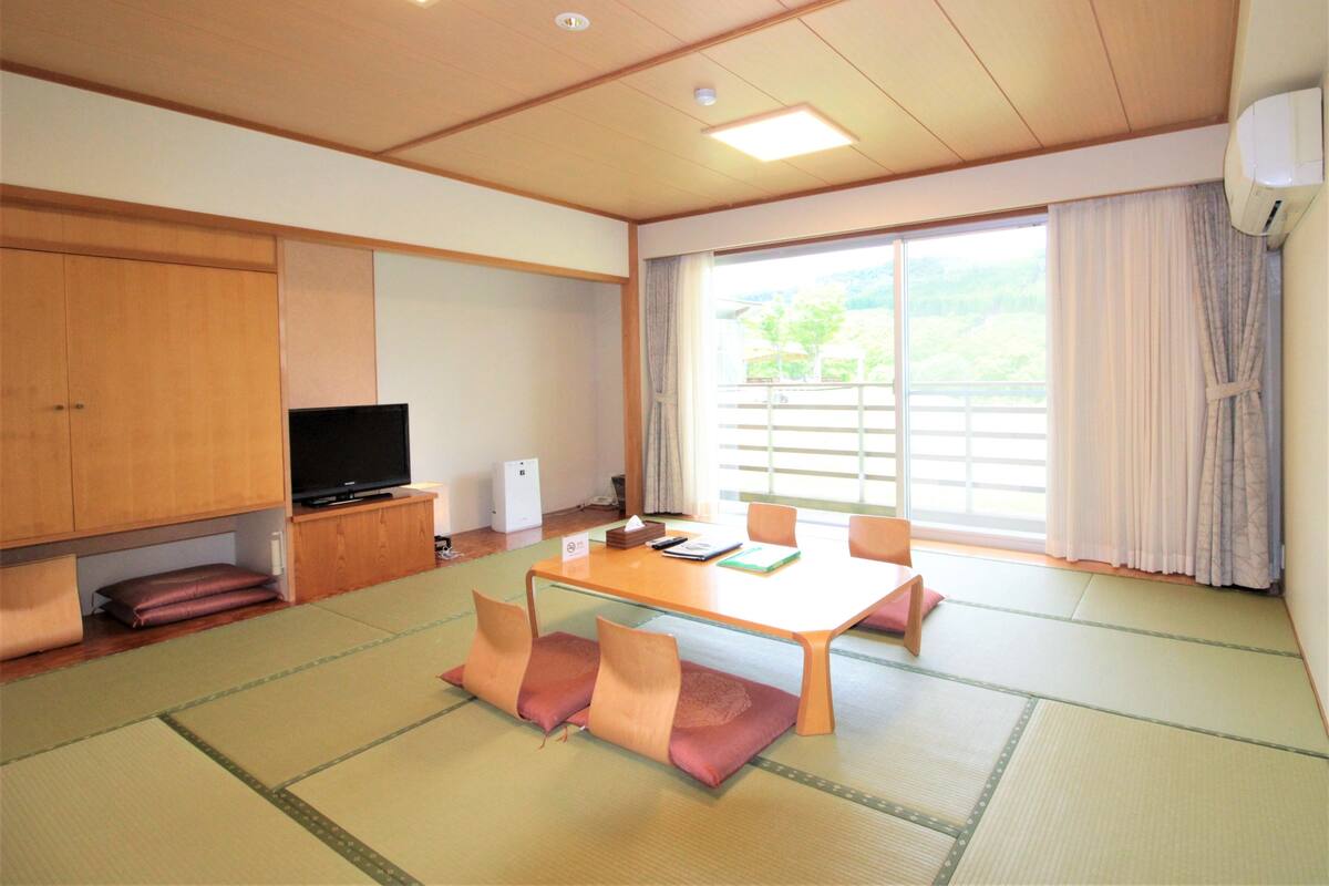Een traditionele Japanse kamer met tatami vloer, een lage tafel met kussens, een televisie en een schuifdeur die leidt naar een balkon met uitzicht op groen.