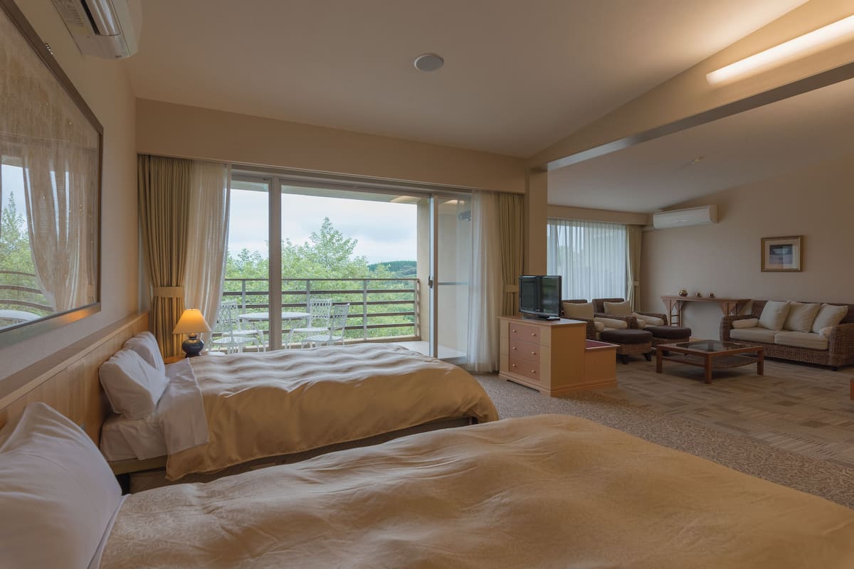 Een hotelkamer met twee bedden, een televisie, een bank en een balkon met uitzicht op groen.