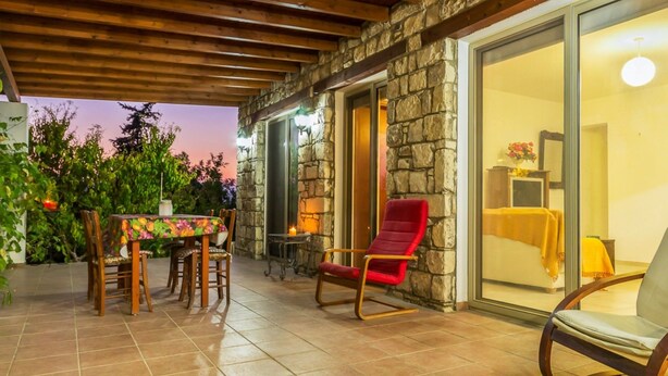 Un patio con pared de piedra, una mesa de comedor con sillas, una silla roja y vistas hacia una habitación con cama y mesita de noche.