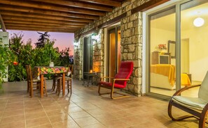 A patio with a stone wall, a dining table with chairs, a red chair, and a view into a room with a bed and a nightstand. - The Lemon Tree Lodge Rustic Village (Amargeti)