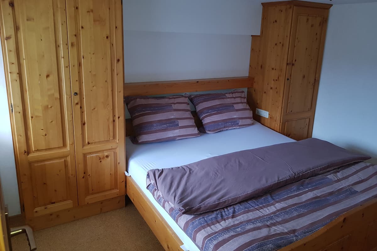 A bedroom with a wooden bed frame, a white mattress, and two striped pillows. There is a wooden wardrobe on one side and a ceiling light fixture.