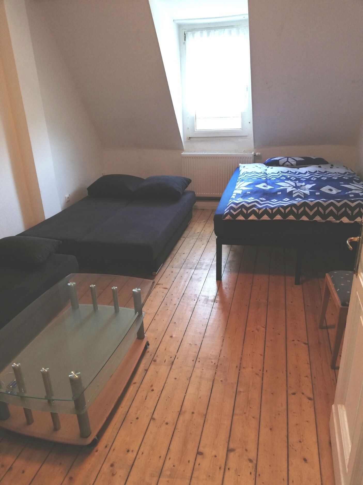 Ein kleiner, gemütlicher Raum mit schrägem Dach, einem Bett mit gemustertem Überwurf, einem kleinen Tisch mit Glasplatte und einem Fenster, das natürliches Licht hereinlässt.