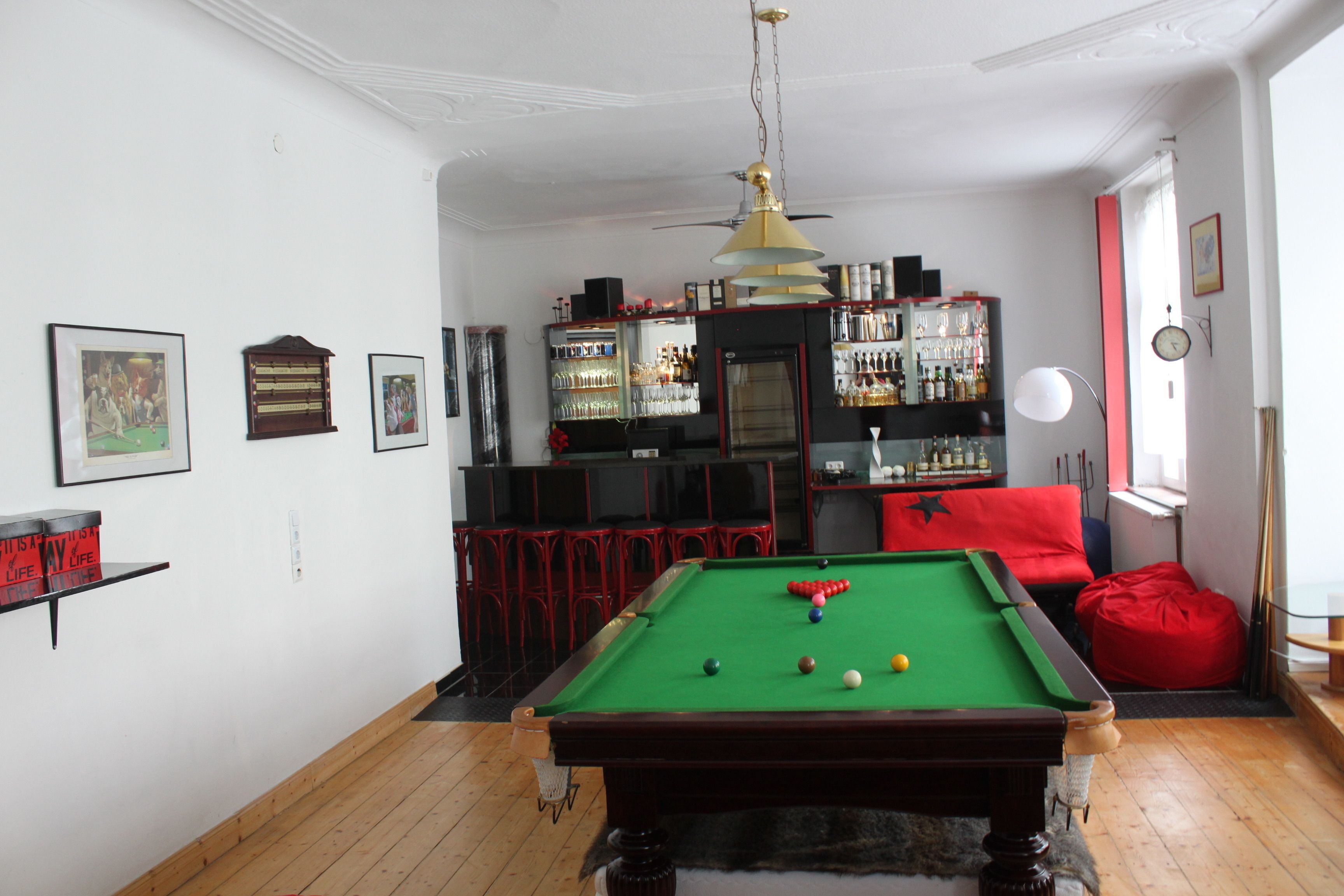 Una mesa de billar en una sala con zona de bar, un sofá rojo y un reloj en la pared.