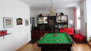 Una mesa de billar en una sala con zona de bar, un sofá rojo y un reloj en la pared.