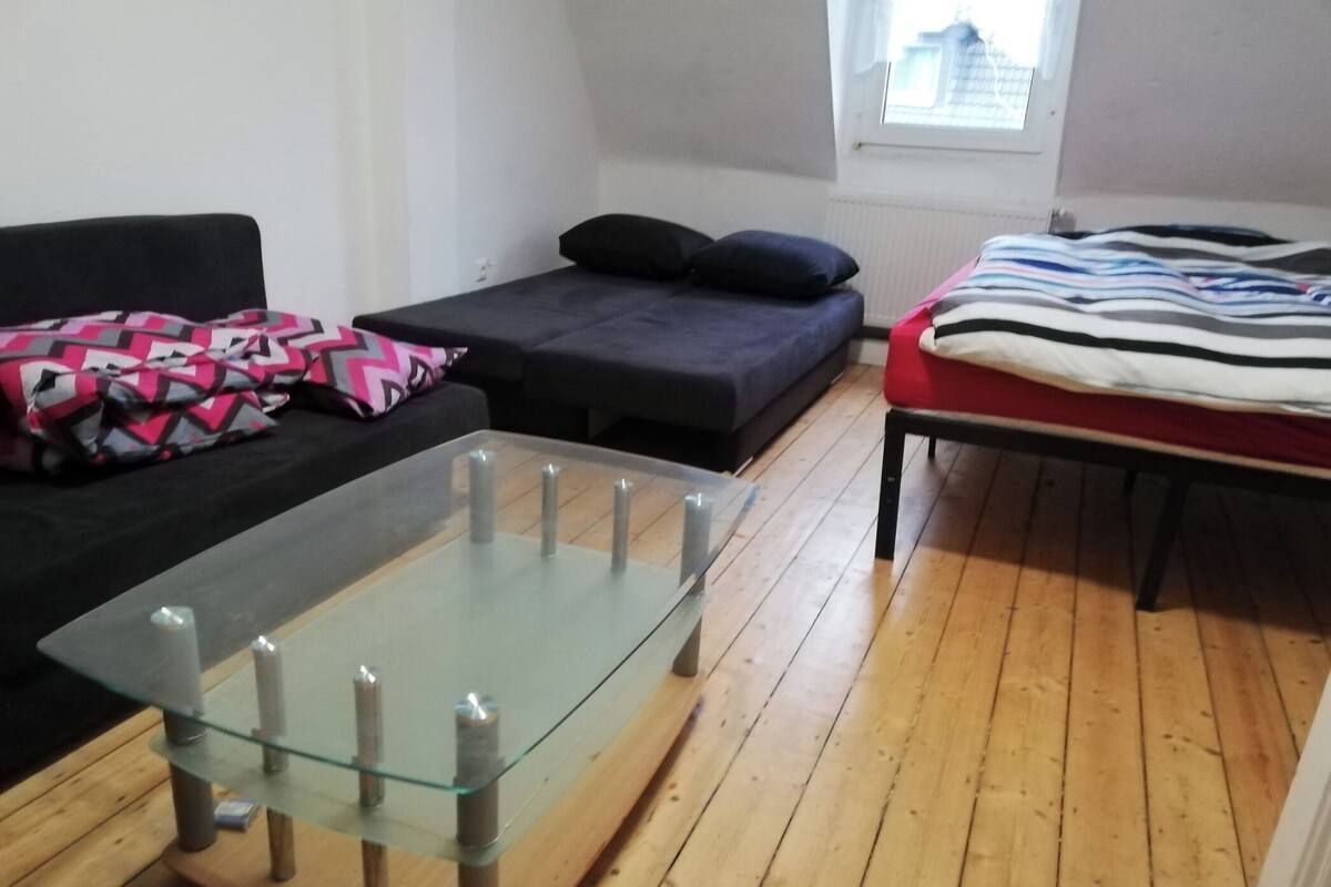 Um espaço de estar compacto com uma mesa de centro de vidro, um sofá com almofadas estampadas, uma cama com manta listrada e uma janela com vista.