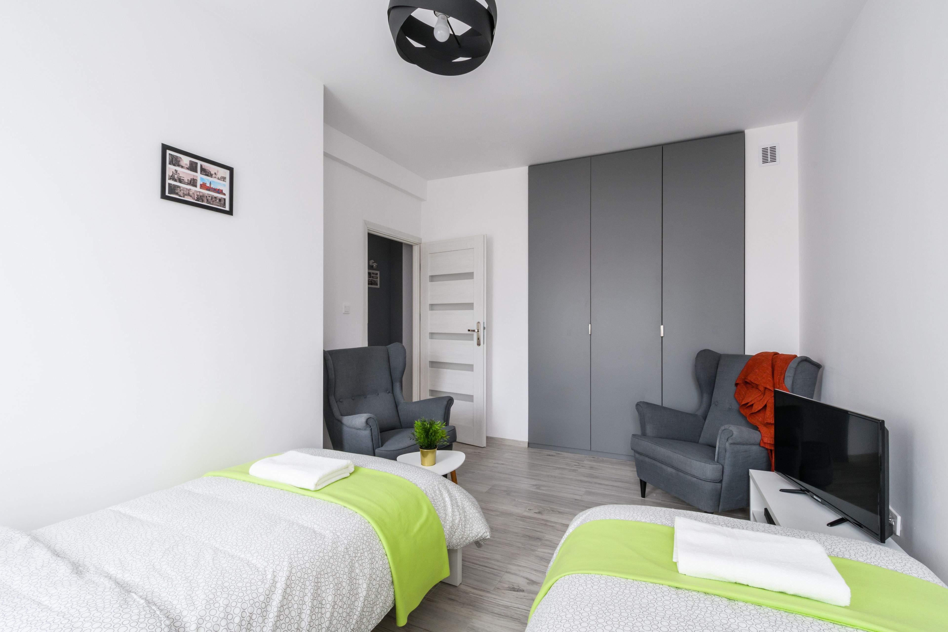 Quarto de hotel moderno com duas camas de solteiro, uma televisão, uma cadeira e uma mesa pequena.