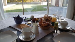 Una mesa de desayuno con cruasanes, tostadas, una cafetera, un vaso de jugo y un bol de fruta.