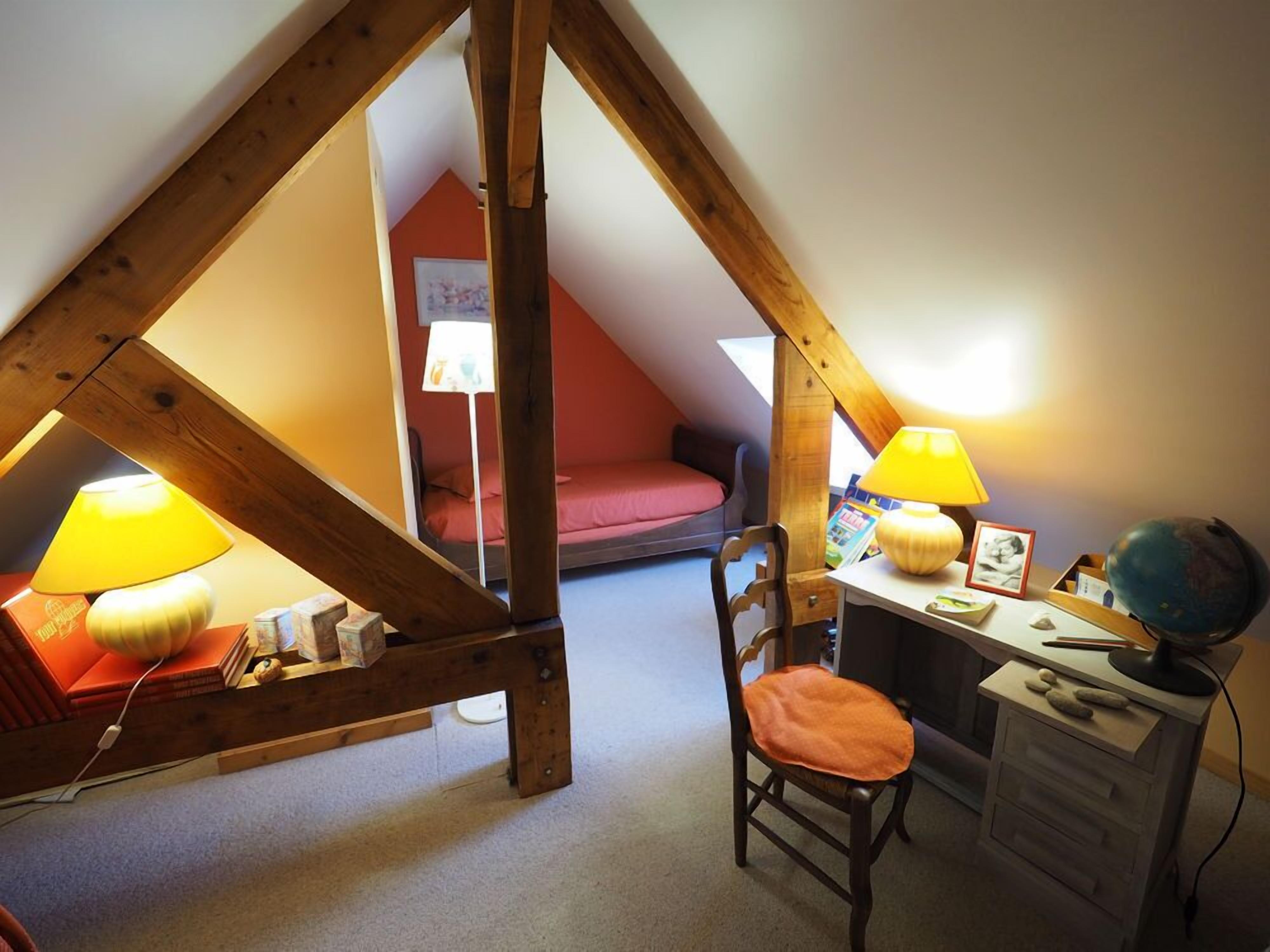 A cozy attic bedroom with a sloped ceiling, wooden beams, a desk with a chair, two lamps, and a globe.