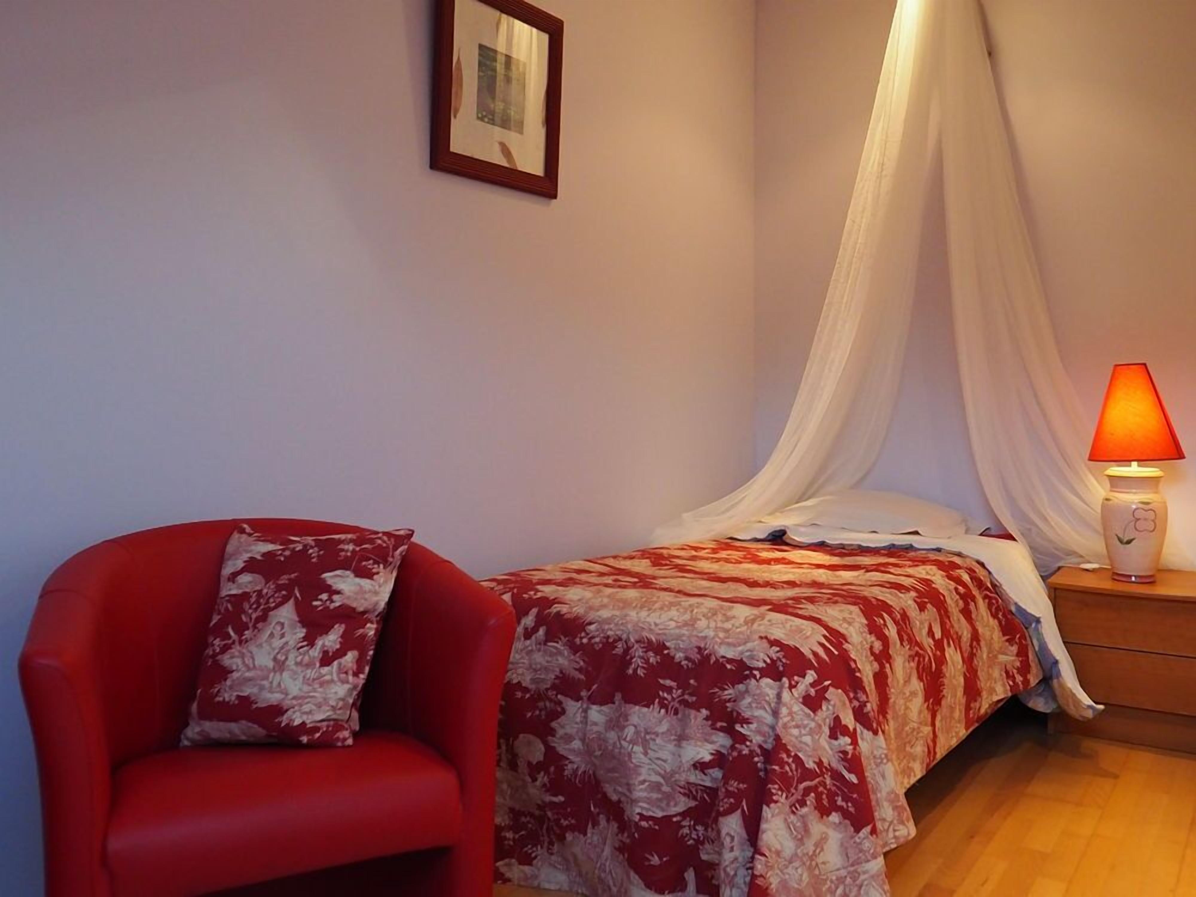 Um quarto com uma cama, uma cadeira vermelha, uma mesa de cabeceira com abajur e um dossel translúcido sobre a cama.
