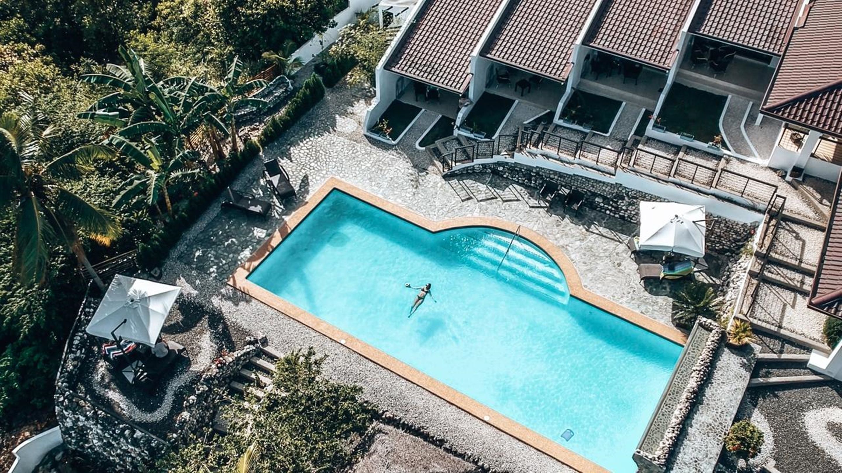 Eine Luftaufnahme eines Resorts mit einem rechteckigen Schwimmbecken, umgeben von Liegestühlen und Sonnenschirmen, einem überdachten Außenbereich mit einem Ziegeldach und üppiger tropischer Vegetation.