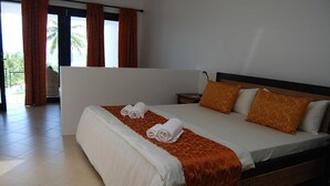 A hotel room with a large bed, two pillows, a nightstand, and a view of palm trees through the window.