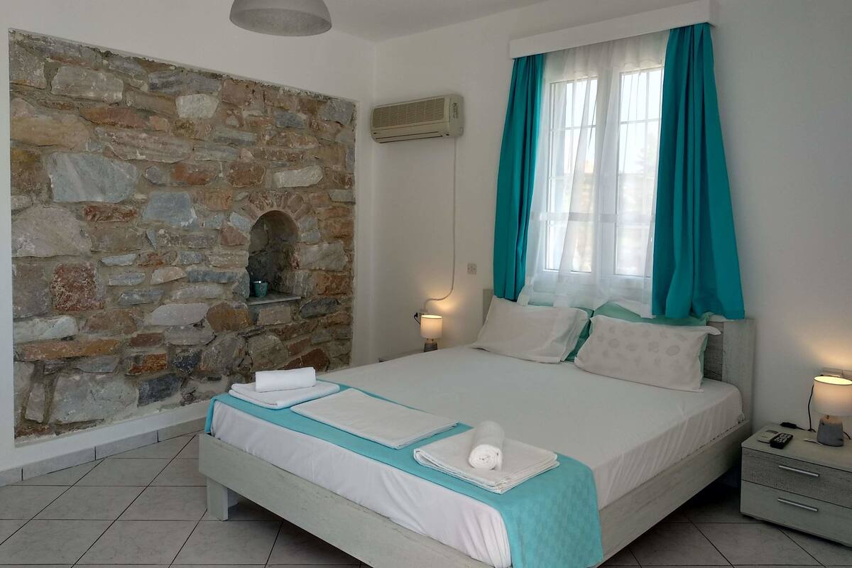Une chambre avec un mur en pierre, un lit avec une literie blanche et des touches de couleur turquoise, une table de chevet avec une lampe, et une fenêtre avec des rideaux turquoise.
